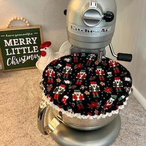May include: A silver KitchenAid Professional 600 stand mixer with a black and white Christmas-themed cover featuring nutcracker figures and poinsettias. The cover has a white lace trim. A green sign reads "Merry Little Christmas."
