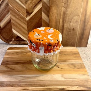 Op de afbeelding: Een heldere glazen pot met een Halloween-thema stoffen deksel. Het deksel is oranje met een patroon van pompoenen, vleermuizen en manen. De pot heeft een witte kanten rand rond de rand. De pot staat op een houten snijplank.