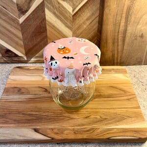 May include: A clear glass jar with a pink fabric cover featuring a Halloween pattern of pumpkins, ghosts, bats, and moons. The cover has a ruffled edge trimmed with white lace and is secured over the top of the jar, which has the word WECK embossed.