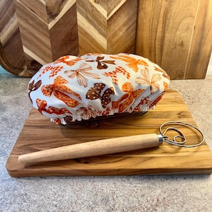 May include: A fabric bowl cover featuring a white background with a repeating pattern of orange, brown, and tan bows, paired with a wooden-handled metal dough whisk. The cover has an elasticized edge and is positioned over a rectangular container.