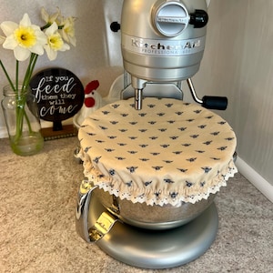 May include: A silver KitchenAid stand mixer with a beige fabric bowl cover featuring a bee pattern. The bowl cover has a white lace trim. The mixer has the text "KitchenAid Professional 600" on the side.