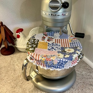 Peut inclure: Un batteur sur socle KitchenAid Professional 600 argenté avec un couvercle de bol patchwork. Le couvercle présente des motifs floraux et géométriques de différentes couleurs, bordés de dentelle blanche. Une figurine de coq décorative est visible en arrière-plan.