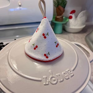 Op de afbeelding: Een witte pannendekselhoes met een kersenpatroon. De hoes heeft een rode stippelrand en een lus om op te hangen. De hoes ligt op een witte Lodge-pan.