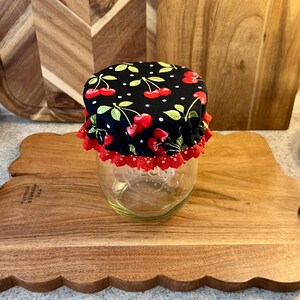 May include: A glass jar with a black cherry-patterned fabric lid and red lace trim. The lid features red cherries, green leaves, and white polka dots on a black background. The jar sits on a wooden cutting board.