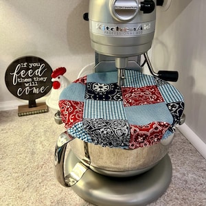 May include: A silver KitchenAid Professional 600 stand mixer with a patchwork bowl cover. The cover features red paisley, navy floral, and blue gingham fabric squares. A decorative sign with the text "If you feed them they will come" is in the background.