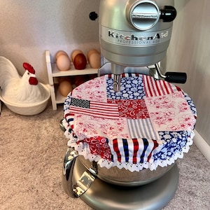 May include: A silver KitchenAid Professional 600 stand mixer with a patriotic patchwork bowl cover featuring an American flag design, red floral patterns, and blue stripes. The cover is trimmed with white lace. A ceramic rooster and eggs are in the background.