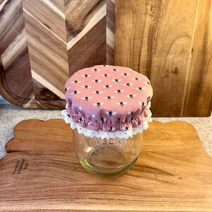 Op de afbeelding: Een heldere glazen pot met een roze stoffen deksel met een patroon van zwarte en gele bijen. De deksel is afgezet met wit kant. De pot staat op een houten snijplank.