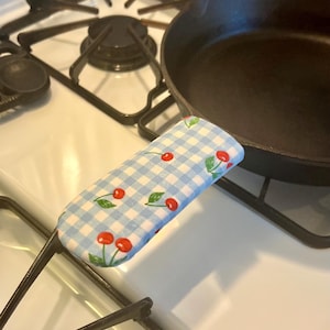 Puede incluir: Una sartén de hierro fundido negro descansa sobre una estufa blanca, junto a un quemador negro. Una funda de mango a cuadros azules y blancos con ilustraciones de cerezas rojas está unida a la sartén.