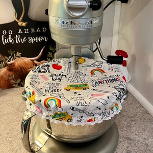Könnte beinhalten: Ein silberner KitchenAid Professional 600 Küchenmixer mit einer weißen, gemusterten Abdeckung. Die Abdeckung zeigt farbenfrohe Illustrationen von Bleistiften, Regenbogen und einem Schulbus mit dem Text "Hey hello" und "You got this!"