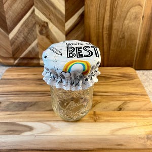May include: A clear glass jar with a fabric lid featuring the text "you're the BEST" above a rainbow design. The lid has a white lace trim. The jar is sitting on a wooden cutting board.