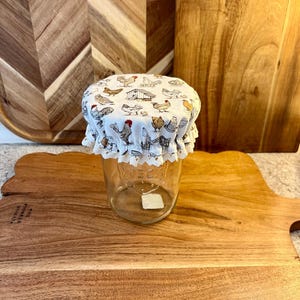 May include: A clear glass jar with a white fabric lid featuring a chicken print. The lid has a ruffled lace trim. The jar is sitting on a wooden cutting board with a scalloped edge. The background includes other wooden cutting boards.