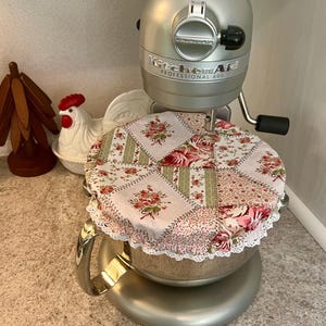 May include: A silver KitchenAid Professional 600 stand mixer with a floral patchwork bowl cover. The cover features pink and green floral patterns with a white lace trim. A ceramic rooster and wooden decor are in the background.