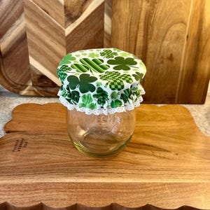 Peut inclure: Un bocal en verre transparent avec un couvercle à motifs de trèfles verts et blancs, bordé de dentelle blanche. Le bocal repose sur une planche à découper en bois. Le couvercle présente divers motifs de trèfles.