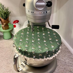 May include: A silver KitchenAid Professional 600 stand mixer with a green cover patterned with yellow and black bees. The cover has a white lace trim. A small rosemary plant and a ceramic rooster are in the background.