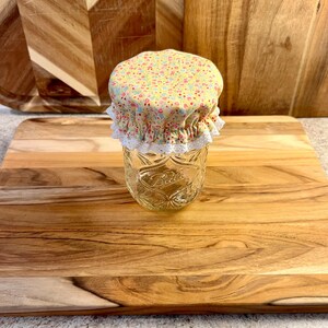 May include: A clear glass jar with a quilted diamond pattern and the embossed word Ball on the front. The jar is topped with a yellow fabric cover featuring a small floral print in pink and blue, finished with a white scalloped lace trim edge.