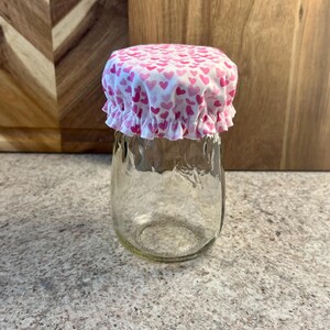May include: A clear glass jar with a pink and white fabric lid with a heart pattern. The lid is elastic and fits snugly over the jar.