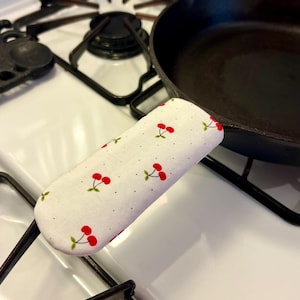 Könnte beinhalten: Ein weißer Griffschutz für eine gusseiserne Pfanne mit einem roten Kirschmuster. Der Bezug befindet sich auf einer schwarzen gusseisernen Pfanne auf einem weißen Herd. Der Griffschutz soll die Hände vor Hitze schützen.