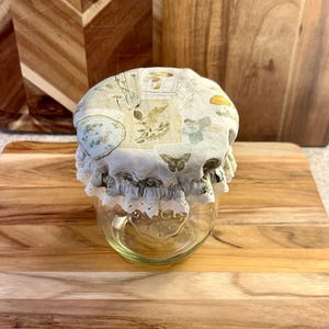 Op de afbeelding: Een heldere glazen pot met een stoffen deksel met een botanische print van paddenstoelen, vlinders en andere natuurlijke elementen. De deksel is afgezet met wit kant. De pot staat op een houten snijplank.