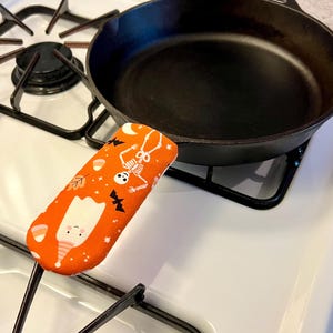 May include: A black cast iron skillet with a fabric handle cover featuring an orange background with a pattern of white ghosts, skeletons, black bats, candy corn, and small leaves. The skillet is positioned on a white stovetop burner grate.