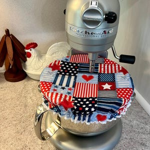 May include: A silver KitchenAid Professional 600 stand mixer with a patriotic patchwork bowl cover. The cover features red, white, and blue fabrics with stars, stripes, and hearts. A white lace trim edges the cover.