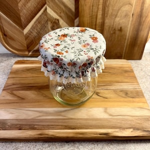 May include: A clear glass jar with a floral fabric lid featuring orange and pink flowers on a white background. The jar is sitting on a wooden chopping board. The jar has the word "WECLY" printed on it.