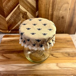 Op de afbeelding: Een heldere glazen pot met een beige stoffen deksel met een patroon van zwarte bijen. De deksel is afgezet met wit kant. De pot staat op een houten snijplank.