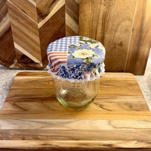 May include: A clear glass jar with a patchwork fabric lid featuring floral, gingham, and striped patterns. The lid is trimmed with white lace. The jar sits on a wooden cutting board, suggesting a kitchen or home decor theme.