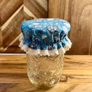 Puede incluir: Una tapa de tarro de tela azul con un borde de encaje blanco y un patrón de vida marina. La tapa está sobre un tarro de vidrio transparente con la palabra "Ball" en relieve en el lateral.