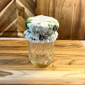 May include: A white fabric jar lid with a green and white floral print and the text "GARDEN" and "PARSLEY". The lid has a white lace trim and is sitting on top of a clear glass jar.