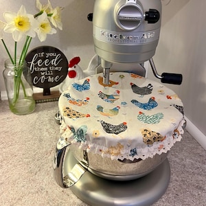 May include: A silver KitchenAid Professional 600 stand mixer with a decorative fabric bowl cover featuring a chicken pattern. The cover has a white lace trim. A vase of daffodils and a decorative sign are in the background.