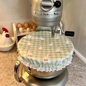 May include: A silver KitchenAid Professional 600 stand mixer with a fabric bowl cover featuring a light green gingham pattern and a repeating white goose print, finished with a delicate white lace trim along the elasticized edge.
