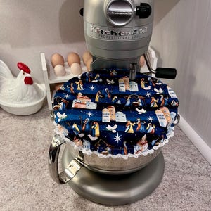 May include: A silver KitchenAid Professional 600 stand mixer with a circular fabric cover placed over the mixing bowl. The cover features a dark blue background with a repeating nativity scene print, white stars, and a white lace trim edge.