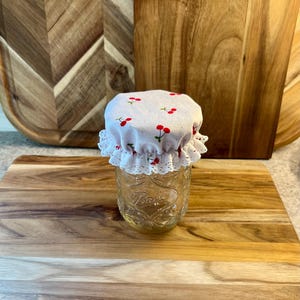 May include: A white fabric jar lid with a red cherry print and white lace trim. The lid is on top of a clear glass jar.