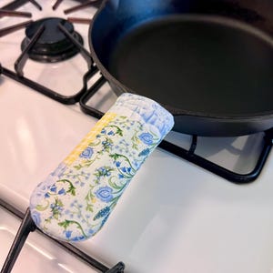 May include: A quilted fabric handle cover featuring a floral pattern with blue flowers and green leaves on a white background, accented by a yellow and white checkered section, fitted onto the handle of a black cast iron skillet on a stove.