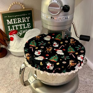 以下が含まれることがあります： サンタクロース、雪だるま、トナカイ、クリスマスツリーの模様が施された、黒いクリスマスをテーマにしたミキサーカバー。カバーには白いレースのトリムが付いており、シルバーのKitchenAidミキサーの上に置かれています。「- have yourself a - MERRY LITTLE Christmas」というテキストの緑色のサインが見えます。