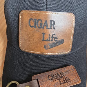 以下が含まれることがあります： 「CIGAR Life」の文字と葉巻のグラフィックが入った茶色の革製パッチが付いた黒いトラッカーハット。同じテキストが入ったお揃いの木製キーホルダーも写っています。