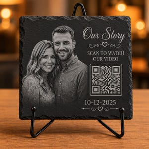 May include: A rectangular black slate plaque featuring a laser-engraved photograph of a couple, the text Our Story, SCAN TO WATCH OUR VIDEO, and the date 10-12-2025. A QR code is centered on the right side, and the plaque rests on a black metal stand.