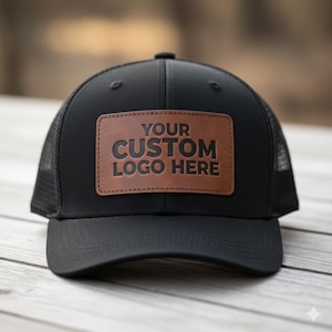 May include: Black trucker hat with a brown leather patch. The patch features the text "YOUR CUSTOM LOGO HERE" in bold, black letters. The hat has a curved brim and a mesh back.