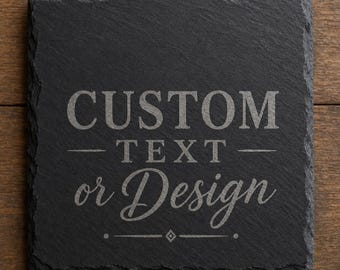 May include: A square black slate coaster with rough, uneven edges. The center features the engraved text CUSTOM, followed by the word TEXT, and the phrase or Design in a script font, finished with a small diamond-shaped decorative element.