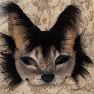 K&ouml;nnte beinhalten: Eine graue und schwarze pelzige Katzenmaske mit einem Spiralmuster auf der Stirn. Die Maske hat schwarzes Fell um die Augen und Ohren.