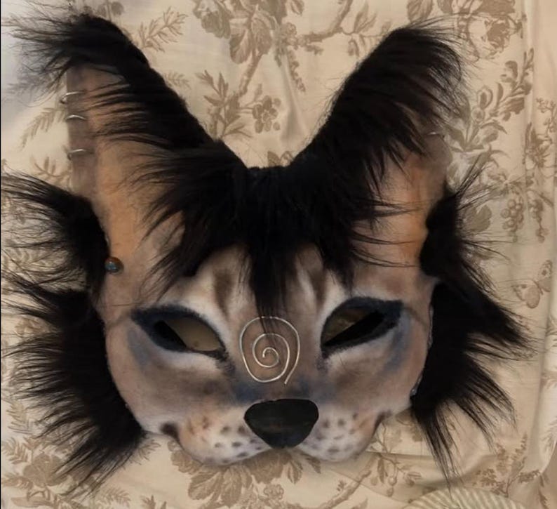 K&ouml;nnte beinhalten: Eine graue und schwarze pelzige Katzenmaske mit einem Spiralmuster auf der Stirn. Die Maske hat schwarzes Fell um die Augen und Ohren.