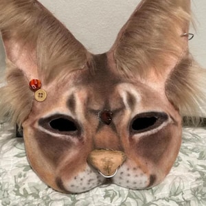 K&ouml;nnte beinhalten: Eine braune und wei&szlig;e Filzkatzenmaske mit gro&szlig;en Ohren, schwarzen Augen und einer kleinen Nase. Die Maske hat einen roten Knopf am linken Ohr und einen braunen Knopf am rechten Ohr.