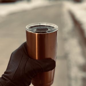 May include: A copper-colored stainless steel travel mug with a black lid. The mug is being held by a gloved hand.