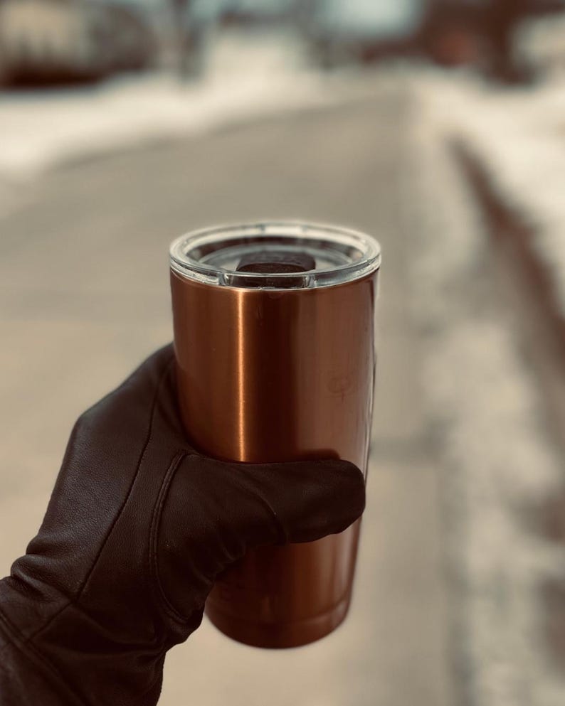 May include: A copper-colored stainless steel travel mug with a black lid. The mug is being held by a gloved hand.