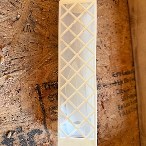 May include: A rectangular, off-white plastic object with a diamond-patterned grid design. The object is long and narrow, with a solid top and a hollow interior. The item is likely a component or part of a larger structure or device.