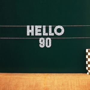May include: A dark green chalkboard with the words "HELLO 90" in silver glitter. Two white lines are drawn across the board. A brown and white checkered mug is on the right side of the image.