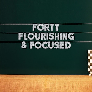 Pode incluir: Uma lousa com o texto "FORTY FLOURISHING & FOCUSED" escrito em letras de glitter prateadas.