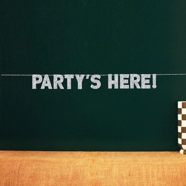 May include: Silver glitter letters spelling "PARTY'S HERE!" hanging on a string against a dark green background.