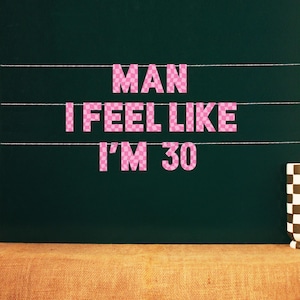 Könnte beinhalten: Rosa karierte Buchstaben schreiben "MAN I FEEL LIKE I'M 30" an einer Schnur vor einem dunkelgrünen Hintergrund. Eine braun-weiß karierte Tasse steht auf einer Jutefläche.