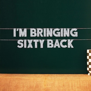 Może przedstawiać: Baner z napisem "I'M BRINGING SIXTY BACK" w srebrnych, brokatowych literach. Baner jest zawieszony na ciemnozielonym tle. Po prawej stronie obrazu widoczny jest brązowo-biały kubek w kratkę.